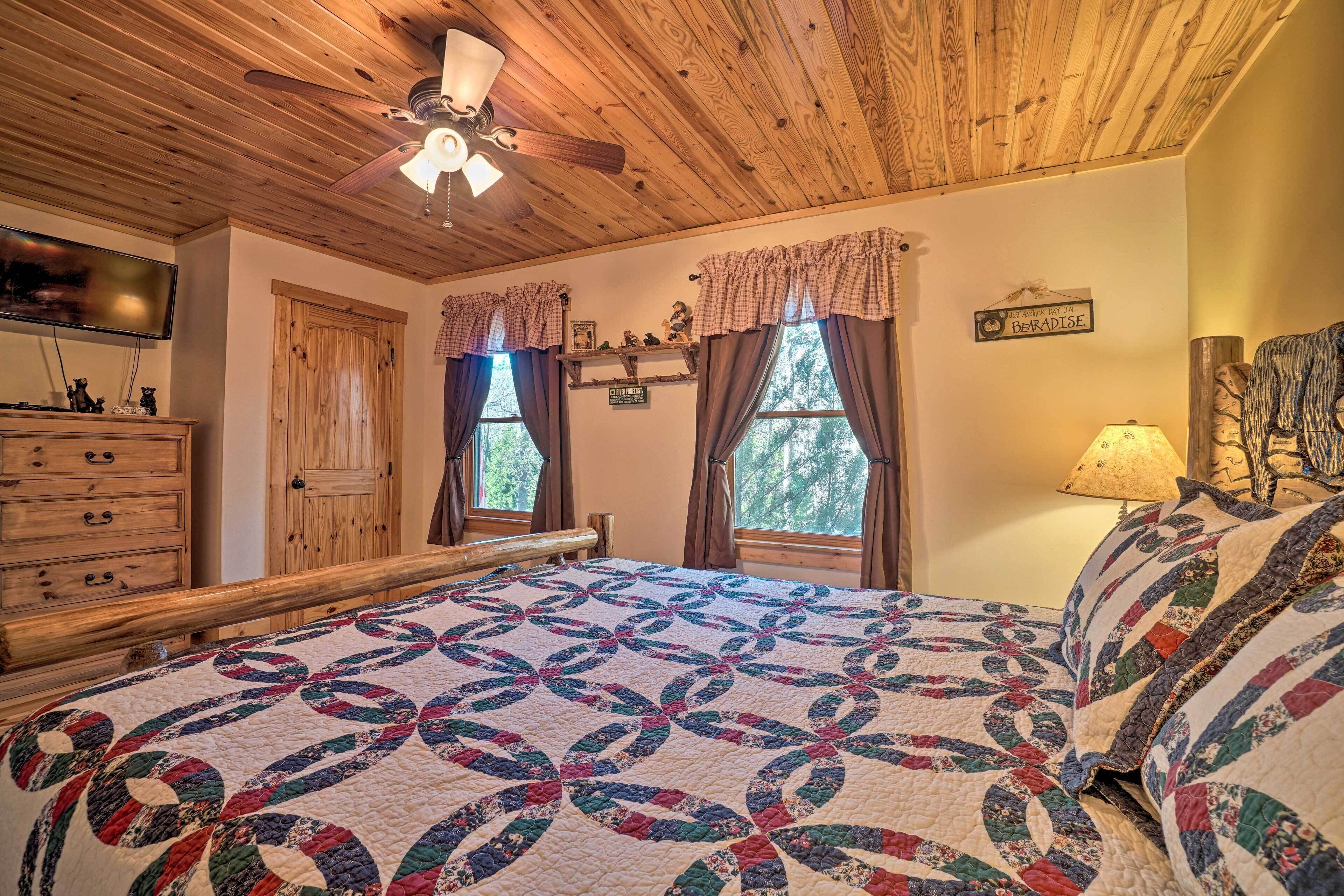 A bedroom with a wooden ceiling, a bed with a colorful quilt, a ceiling fan, and a window with curtains.