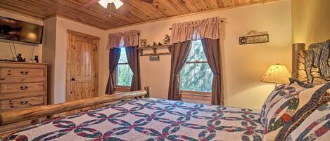 A bedroom with a wooden ceiling, a bed with a colorful quilt, a ceiling fan, and a window with curtains.