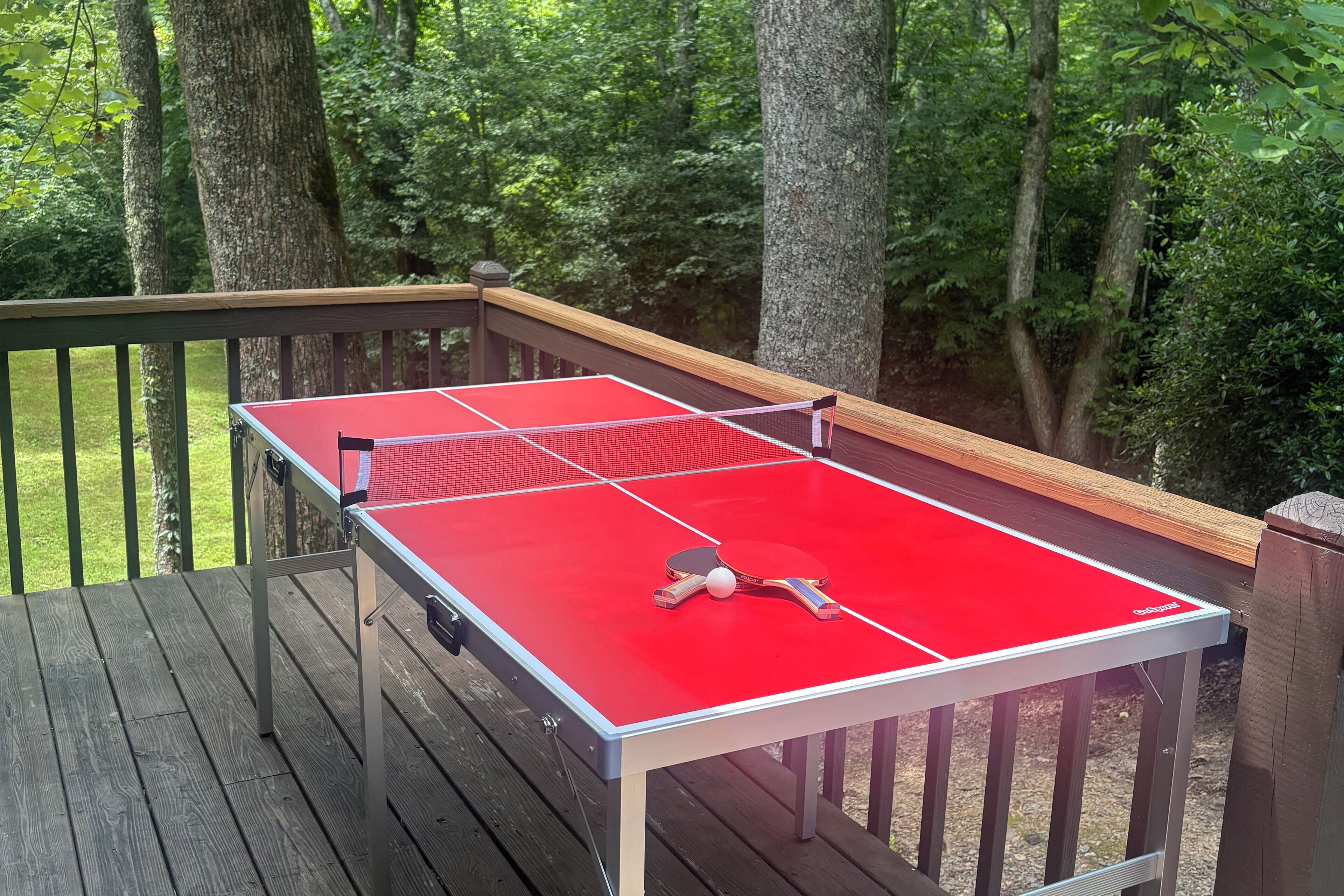 A ping pong table on an outdoor deck with a wooden railing, surrounded by trees.