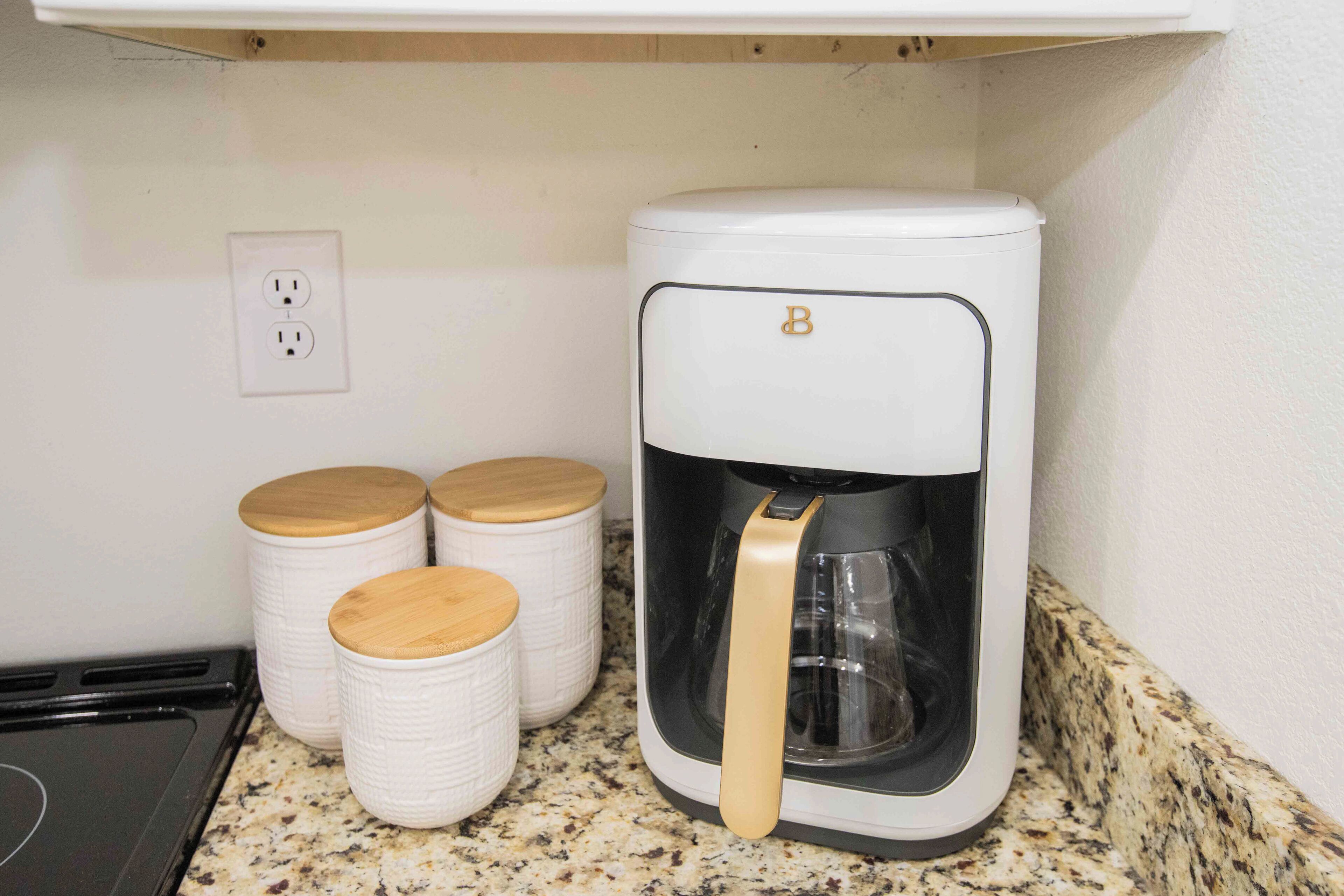 A kitchen with a coffee maker, two white containers with wooden lids, and a power outlet on the wall.