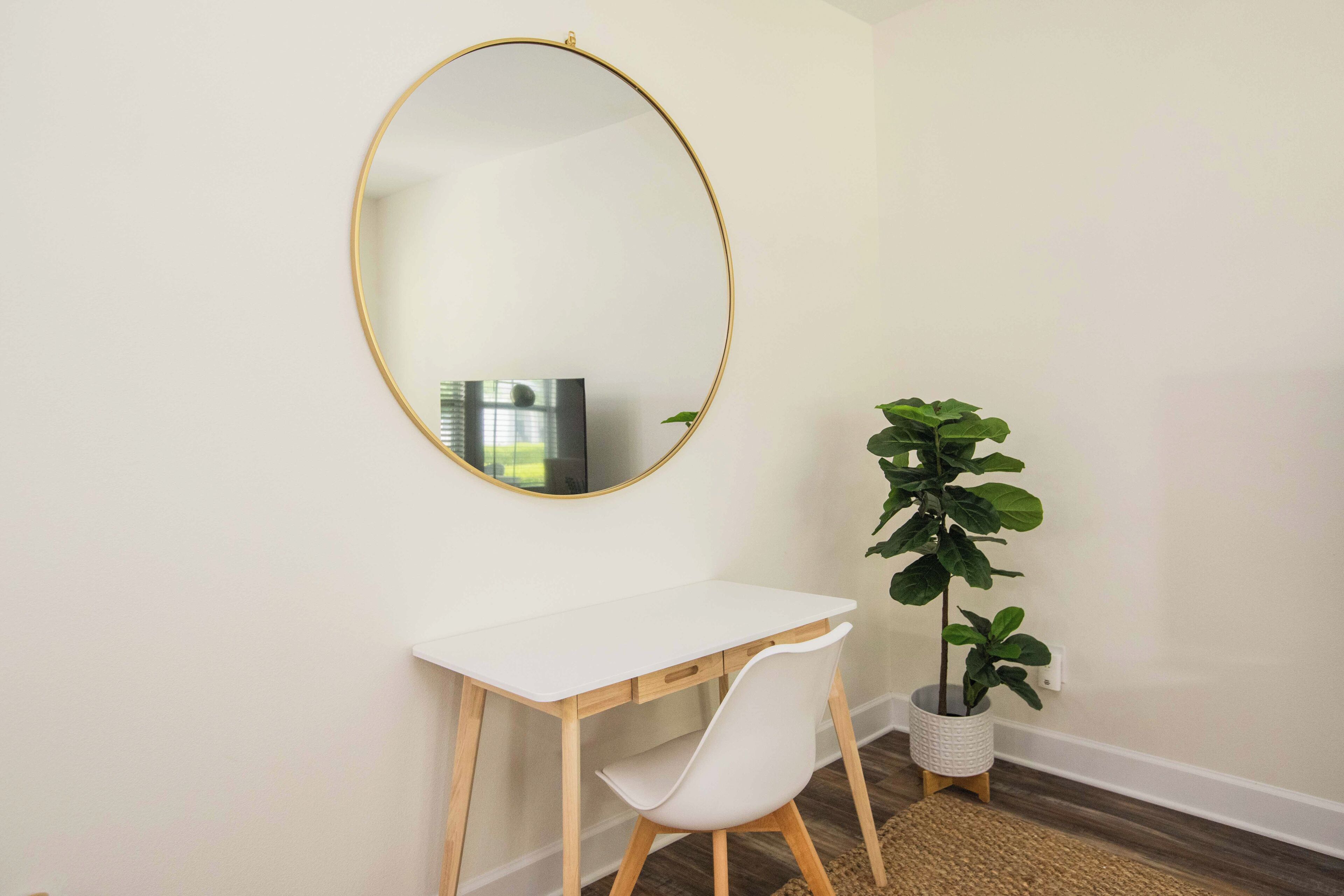 Une petite pièce propre, avec un bureau blanc, un miroir rond et une plante en pot.