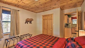 A room with a bed, a wooden ceiling, a window with curtains, a wall-mounted bear decoration, and a kitchen area visible through an open door.