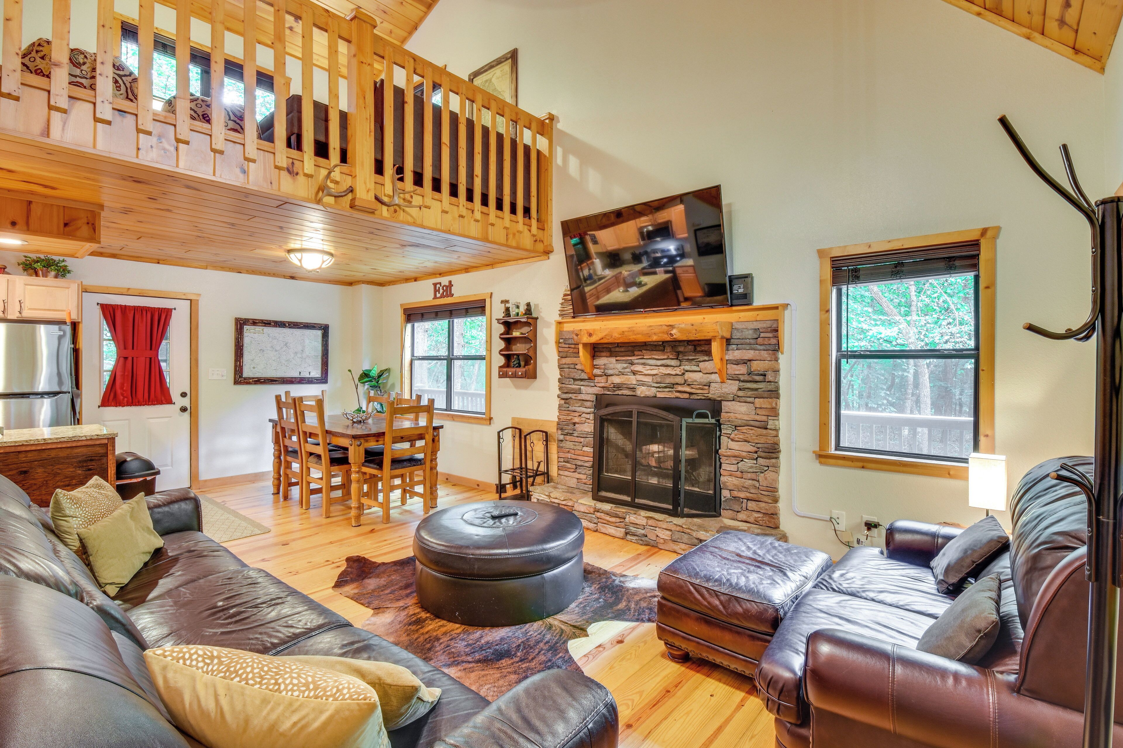 A cozy living room with a stone fireplace, leather sofas, a dining area, and a wooden balcony.
