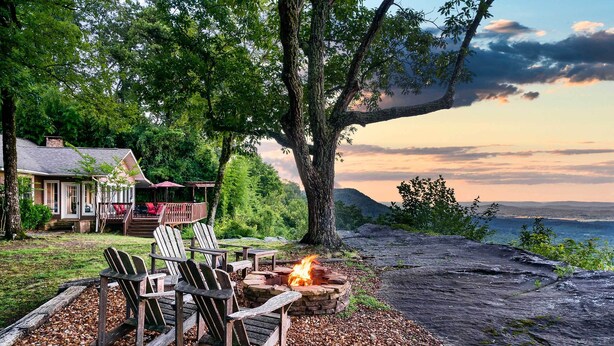 A cozy outdoor seating area with Adirondack chairs and a fire pit, overlooking a scenic view of a lake and mountains.