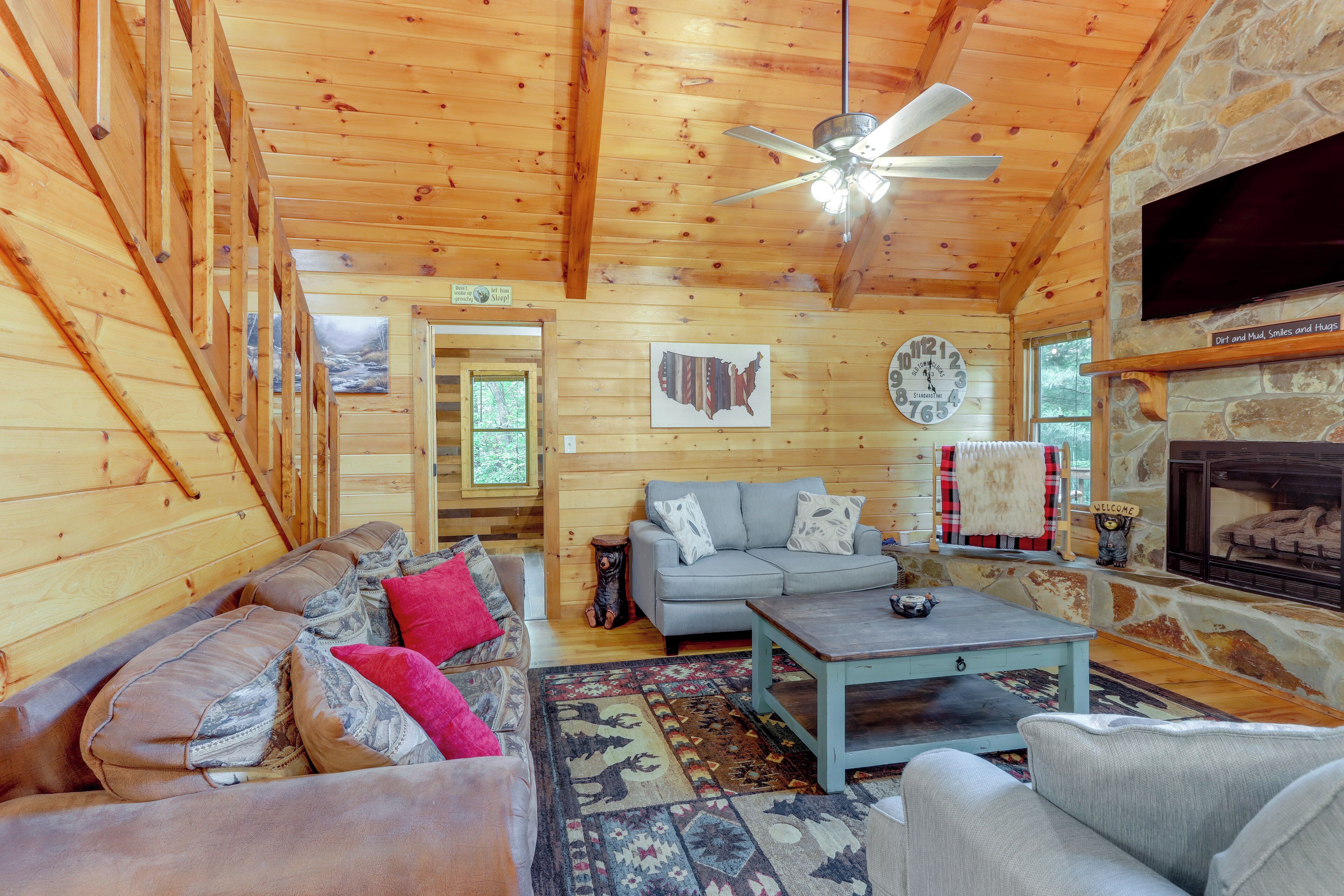 A log cabin interior with a wooden ceiling, a stone fireplace, a gray sofa, a coffee table, and a patterned rug.