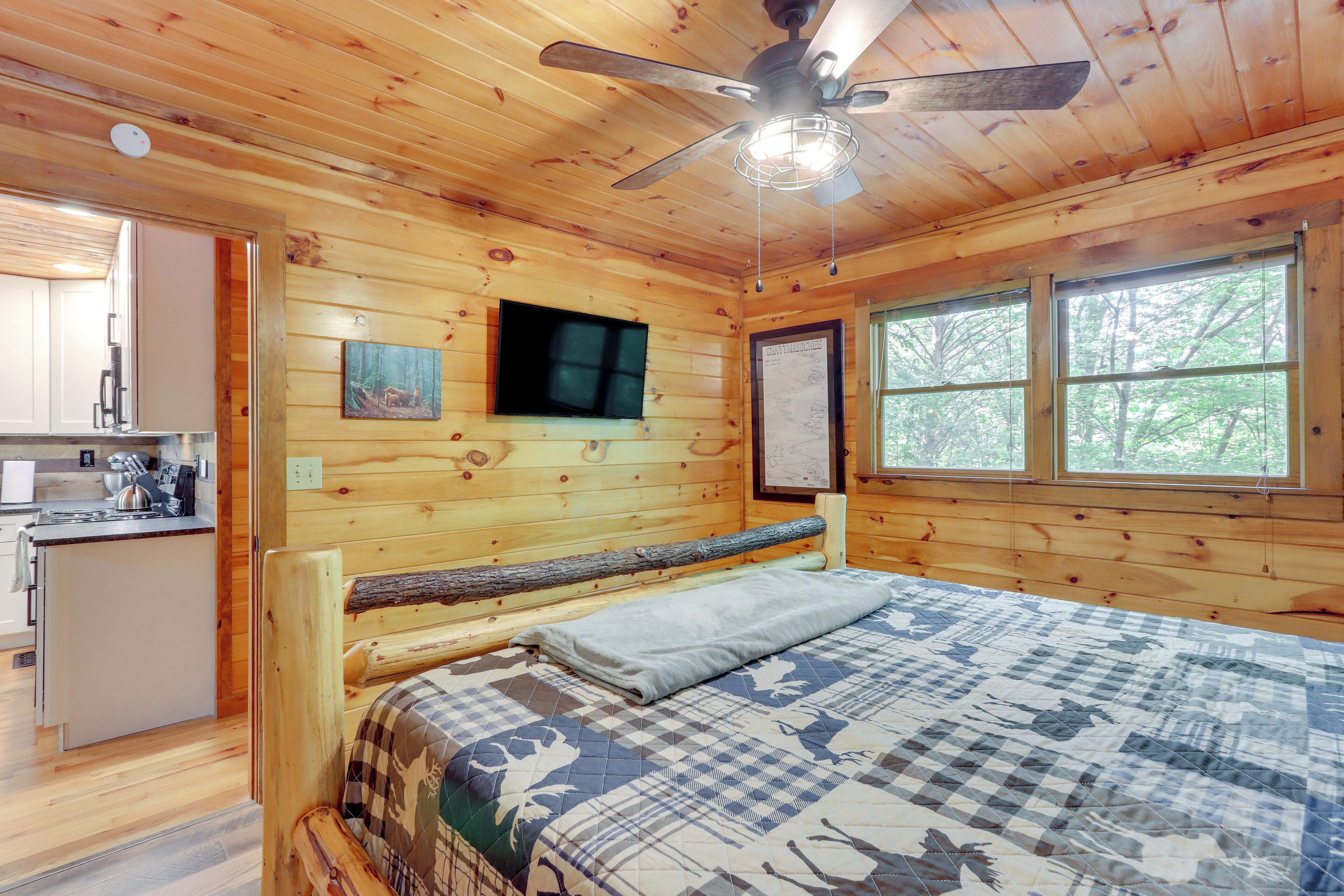 A wooden cabin interior with a bed, a ceiling fan, a television, and a window with a view of trees.