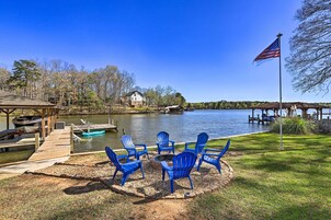 A lakeside deck with blue Adirondack chairs, a fire pit, and a dock with boats.