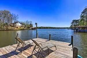 A wooden dock with two lounge chairs facing a calm lake, surrounded by trees and a house in the background.