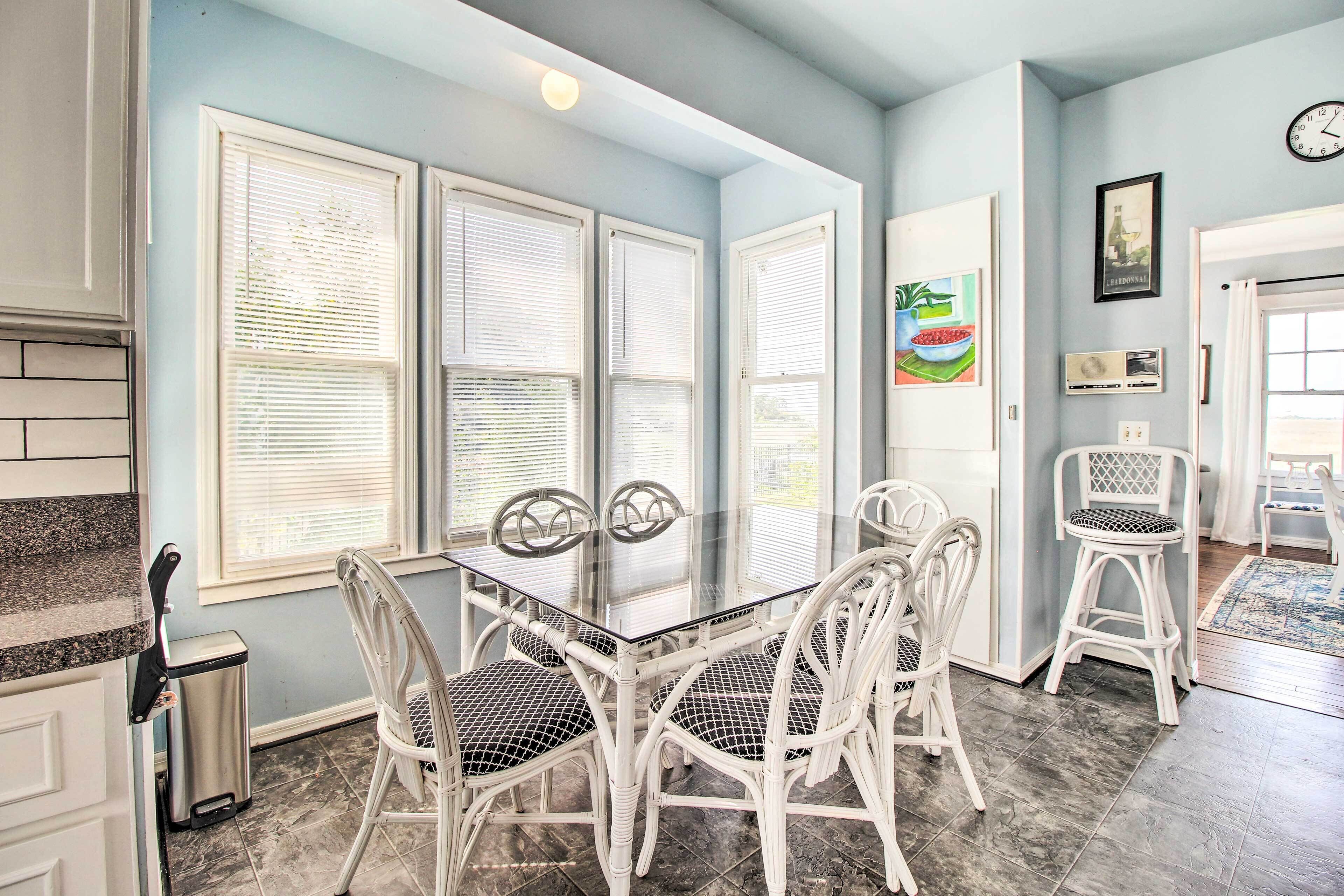 A dining area with a glass-top table surrounded by white wicker chairs, large windows with blinds, and a tiled floor.