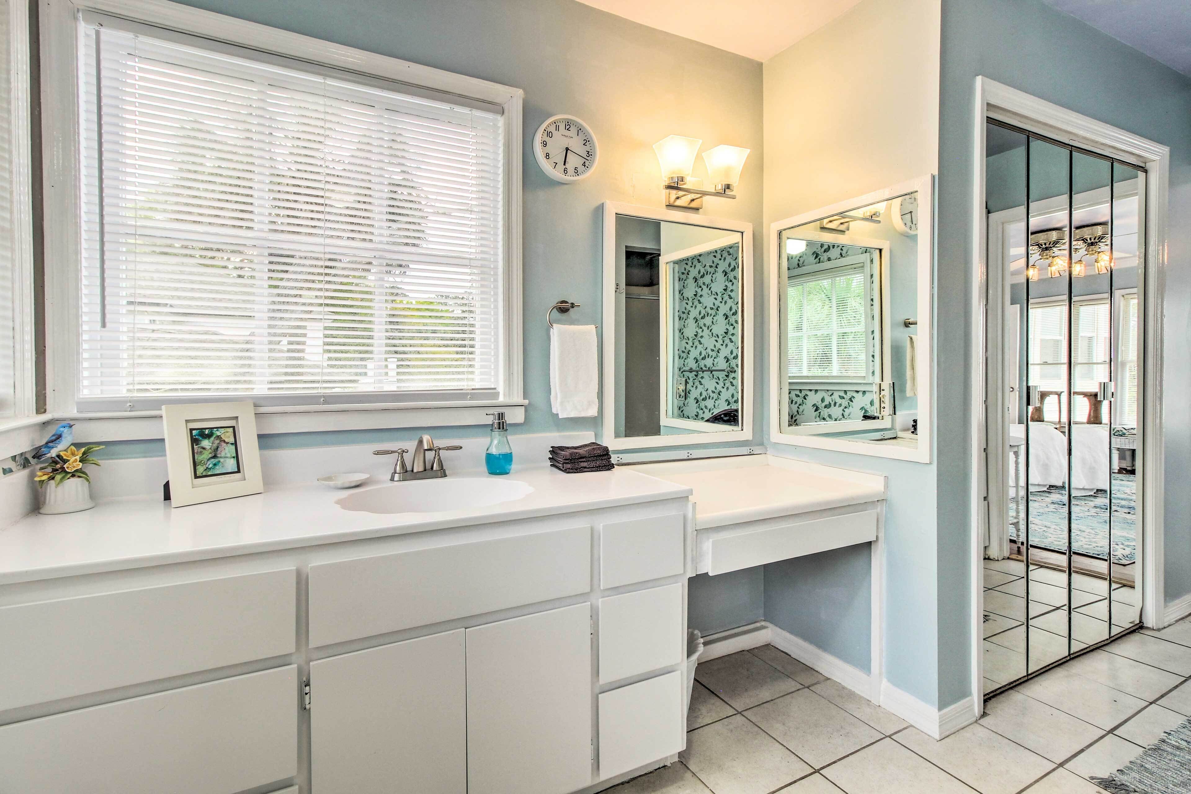 Une salle de bain avec un meuble-lavabo blanc, un grand miroir, une horloge et une fenêtre avec des stores.