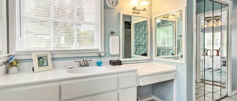 Une salle de bain avec un meuble-lavabo blanc, un grand miroir, une horloge et une fenêtre avec des stores.
