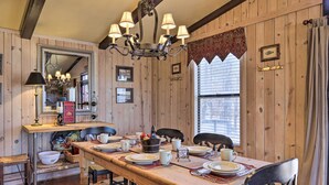 A wooden dining room with a long table set for four, surrounded by chairs. A chandelier hangs above, and a mirror is mounted on the wall.