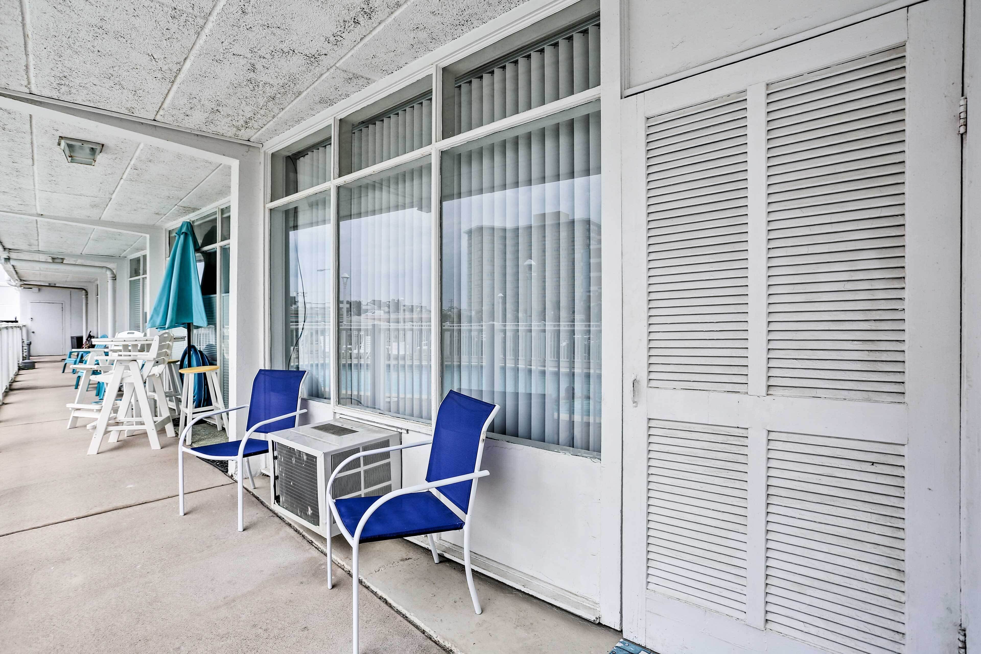 A covered outdoor seating area with white metal chairs and tables, large windows with blinds, and a concrete floor.
