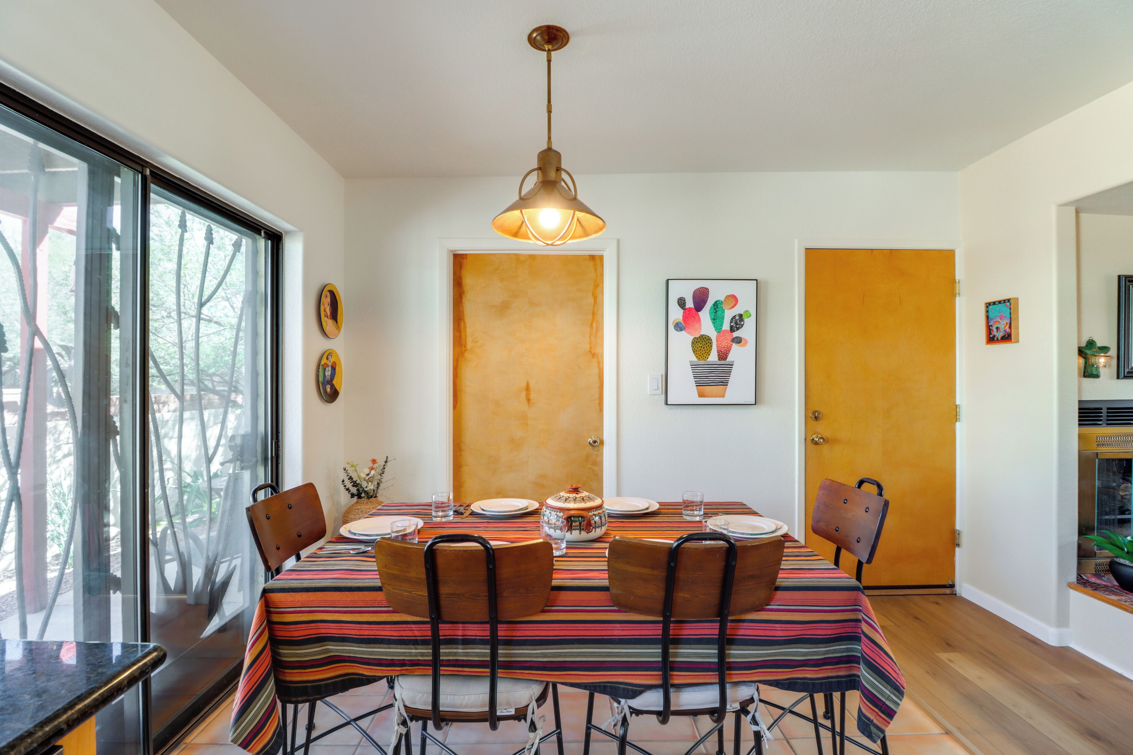 A dining room with a striped tablecloth, wooden chairs, a hanging light fixture, and a door with a fruit painting.