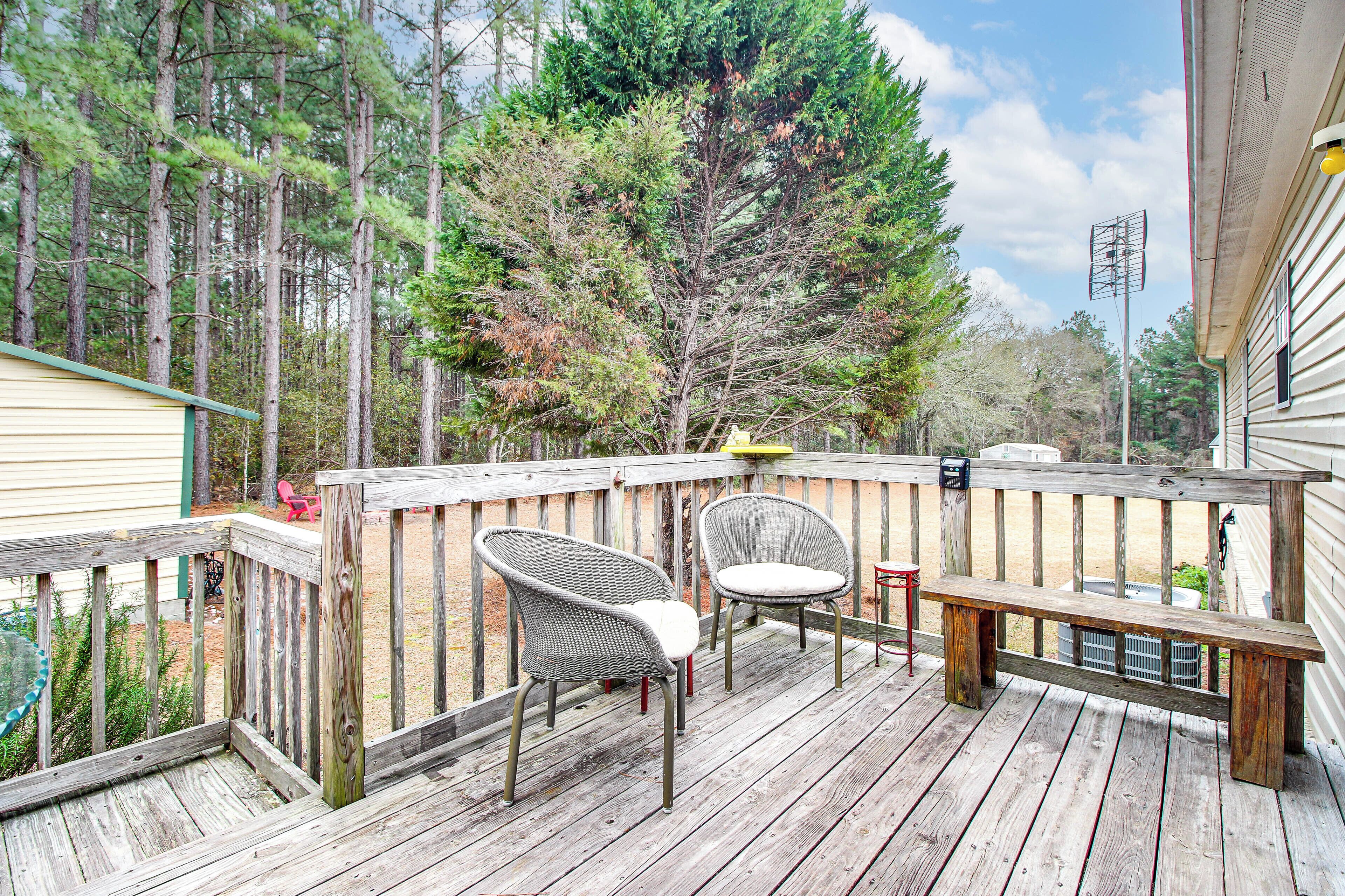A wooden deck with two wicker chairs and a bench, surrounded by trees and a house.