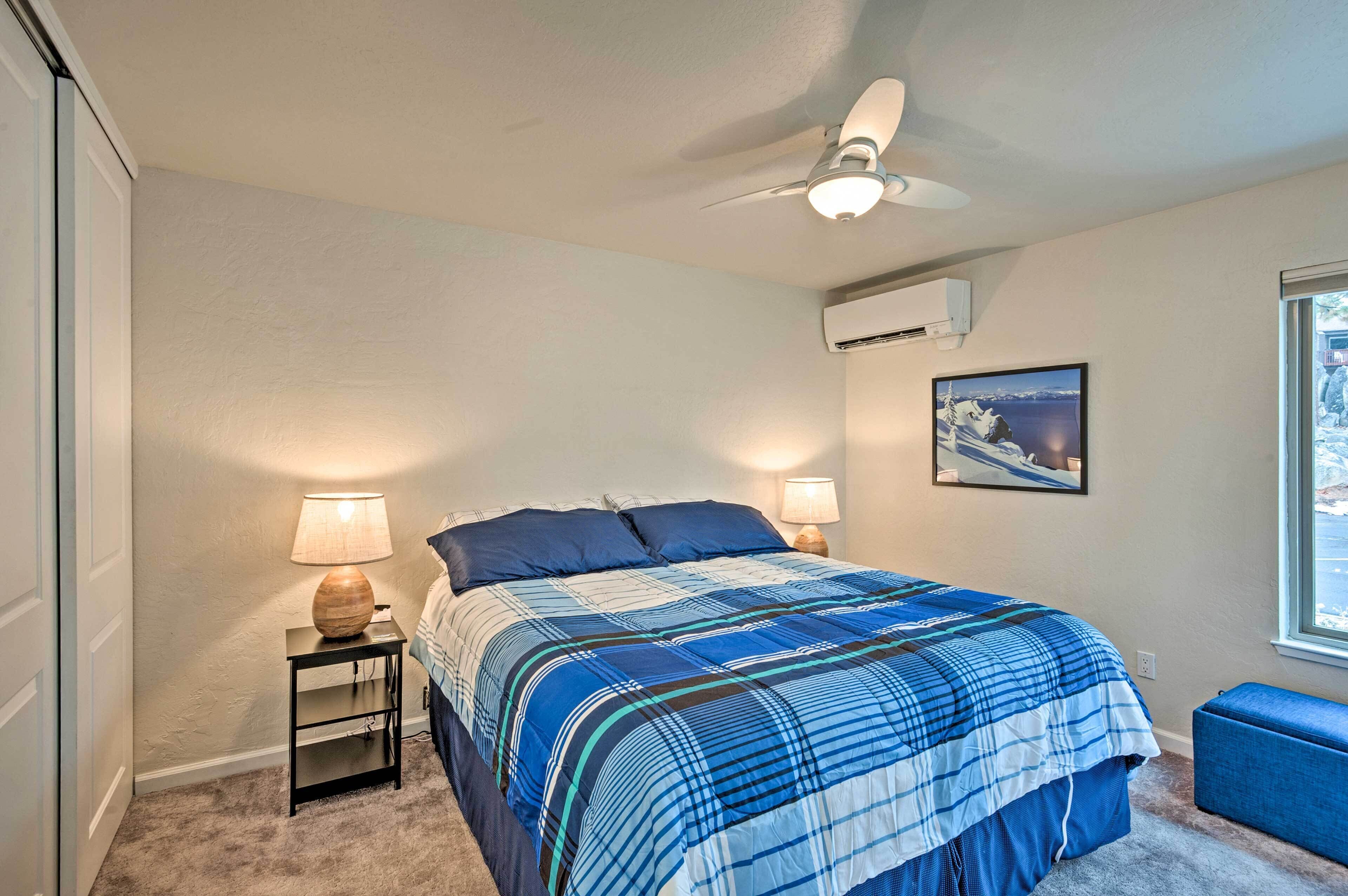 A bedroom with a bed, two bedside lamps, a ceiling fan, an air conditioning unit, a window with a view, and a blue bench.