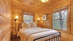 Ein Schlafzimmer einer Holzhütte mit einem Bett, zwei Nachttischlampen, einem Fenster und einem gerahmten Bild an der Wand.