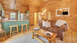 Ein gemütliches Cabin-Interieur mit Holzbalkendecke, einem braunen Ledersofa, einem Holztisch und einer Küchenzeile mit Kühlschrank und Barhocker.
