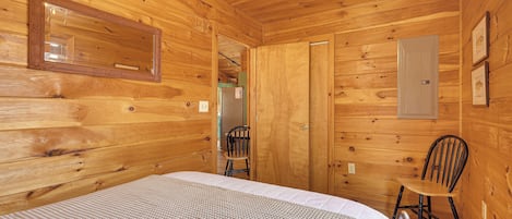 A wooden cabin room with a bed, a chair, and a door leading to another room.