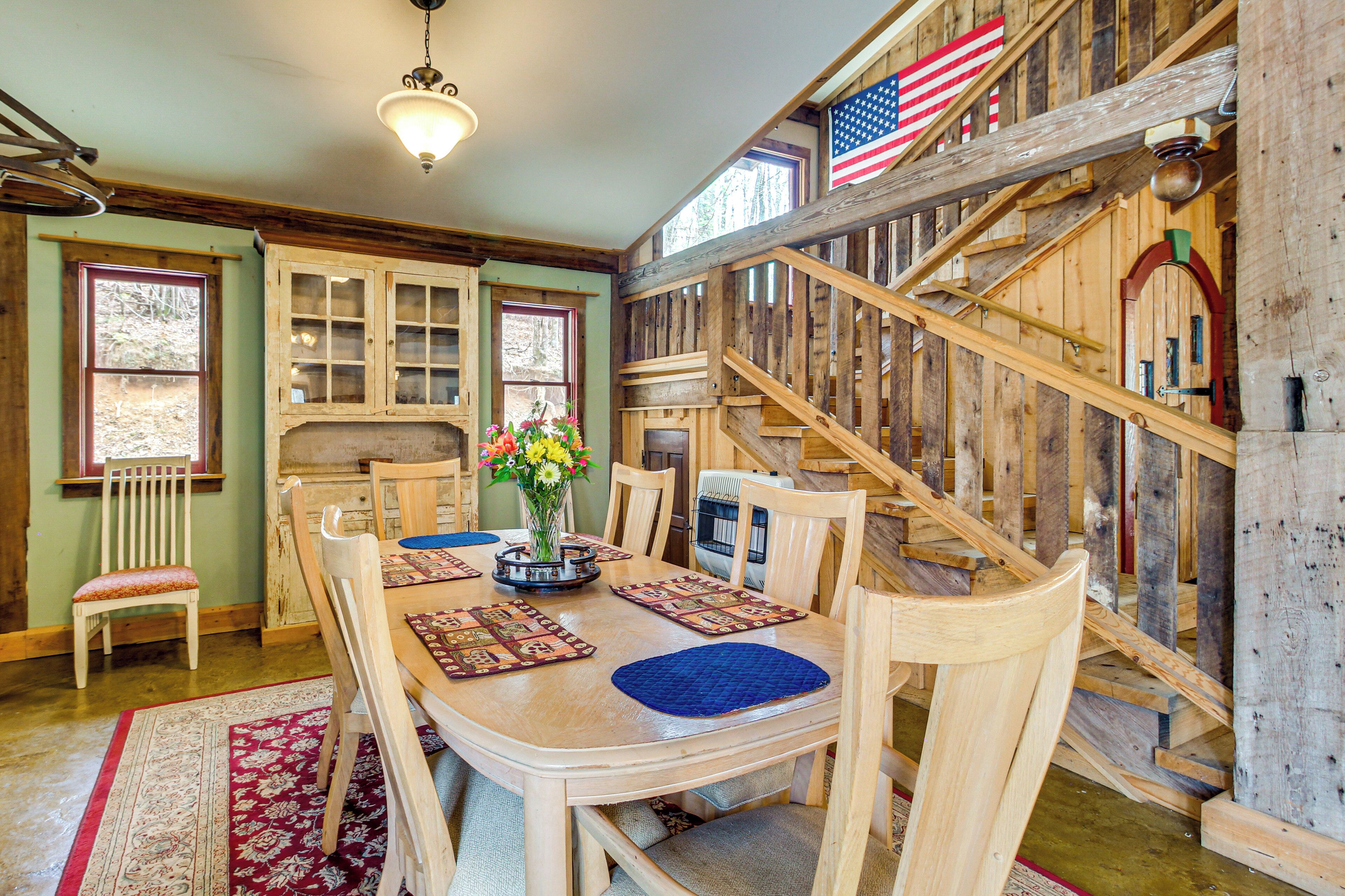 A dining area with a wooden table, chairs, and a vase of flowers. There is a staircase with wooden railings and an American flag.