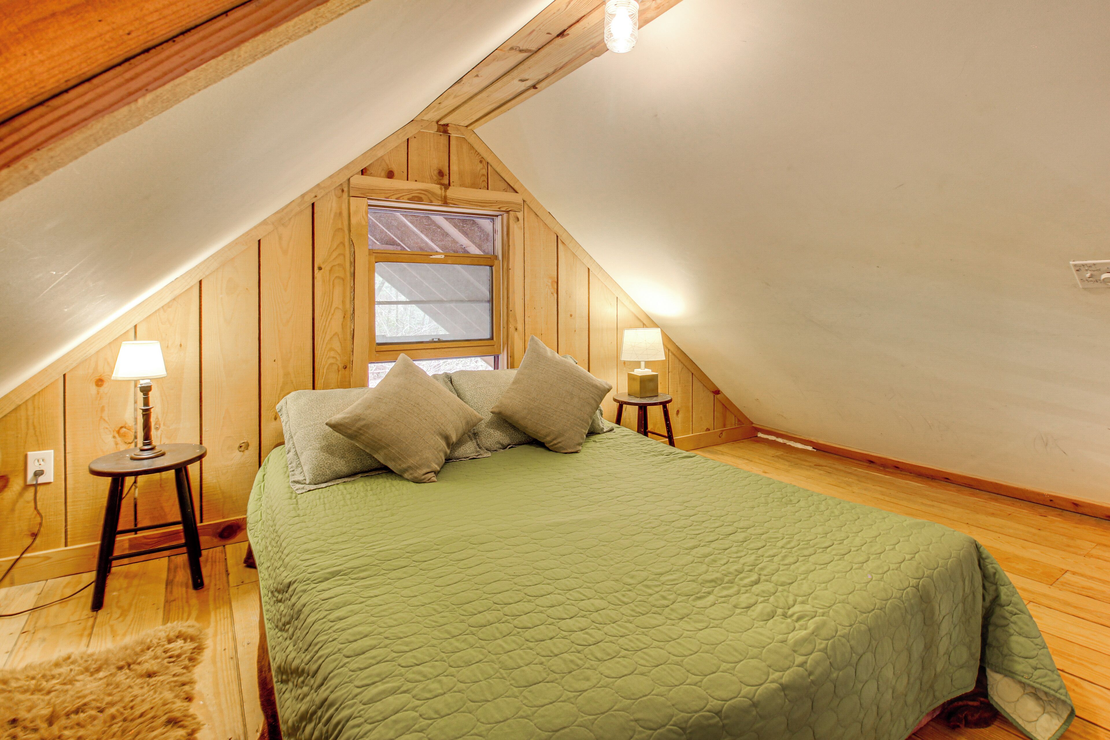 Ein Zimmer mit schrägen Wänden, einem Bett mit grüner Tagesdecke, zwei Nachttischlampen und einem Fenster mit Ausblick.