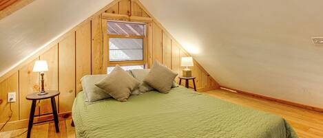 Ein Zimmer mit schrägen Wänden, einem Bett mit grüner Tagesdecke, zwei Nachttischlampen und einem Fenster mit Ausblick.