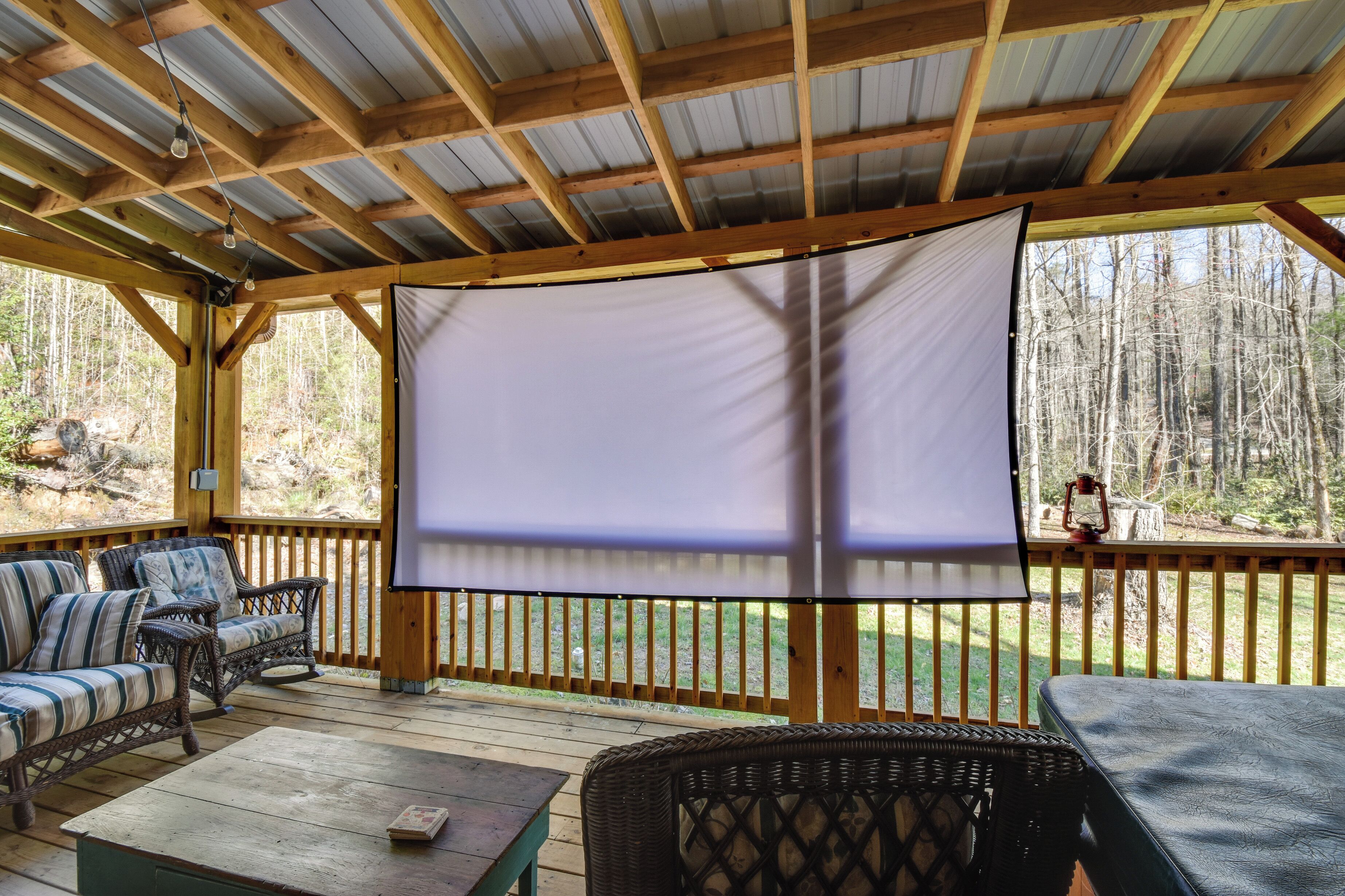 A covered outdoor seating area with a large screen, wooden furniture, and a view of the forest.