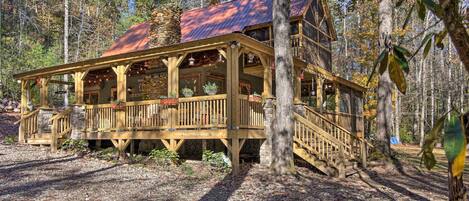 A rustic cabin with a wooden deck, stone foundation, and a metal roof, surrounded by a forest.
