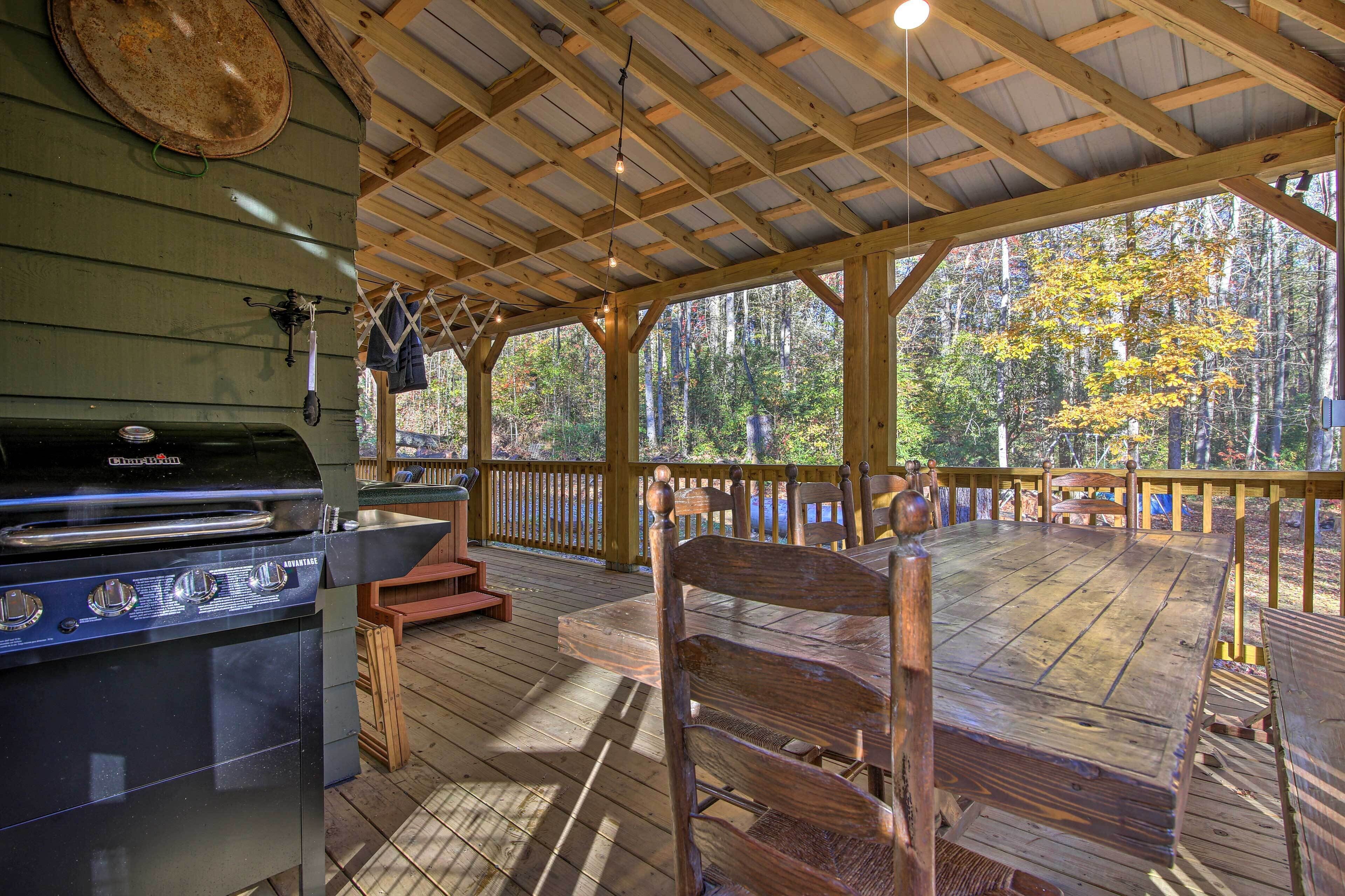 An outdoor covered deck with a grill, wooden furniture, and a view of the forest.