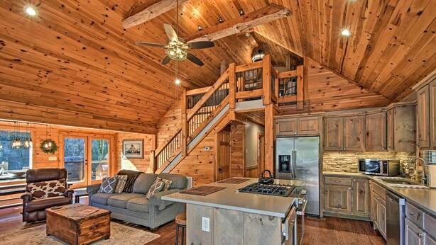 A spacious living area with a large sofa, a coffee table, and a fireplace. There is a kitchen with modern appliances and wooden cabinets. A staircase leads to an upper level. - Blue Ridge Mtns Creekside Cabin w/ Hot Tub + Pier! (Blue Ridge)