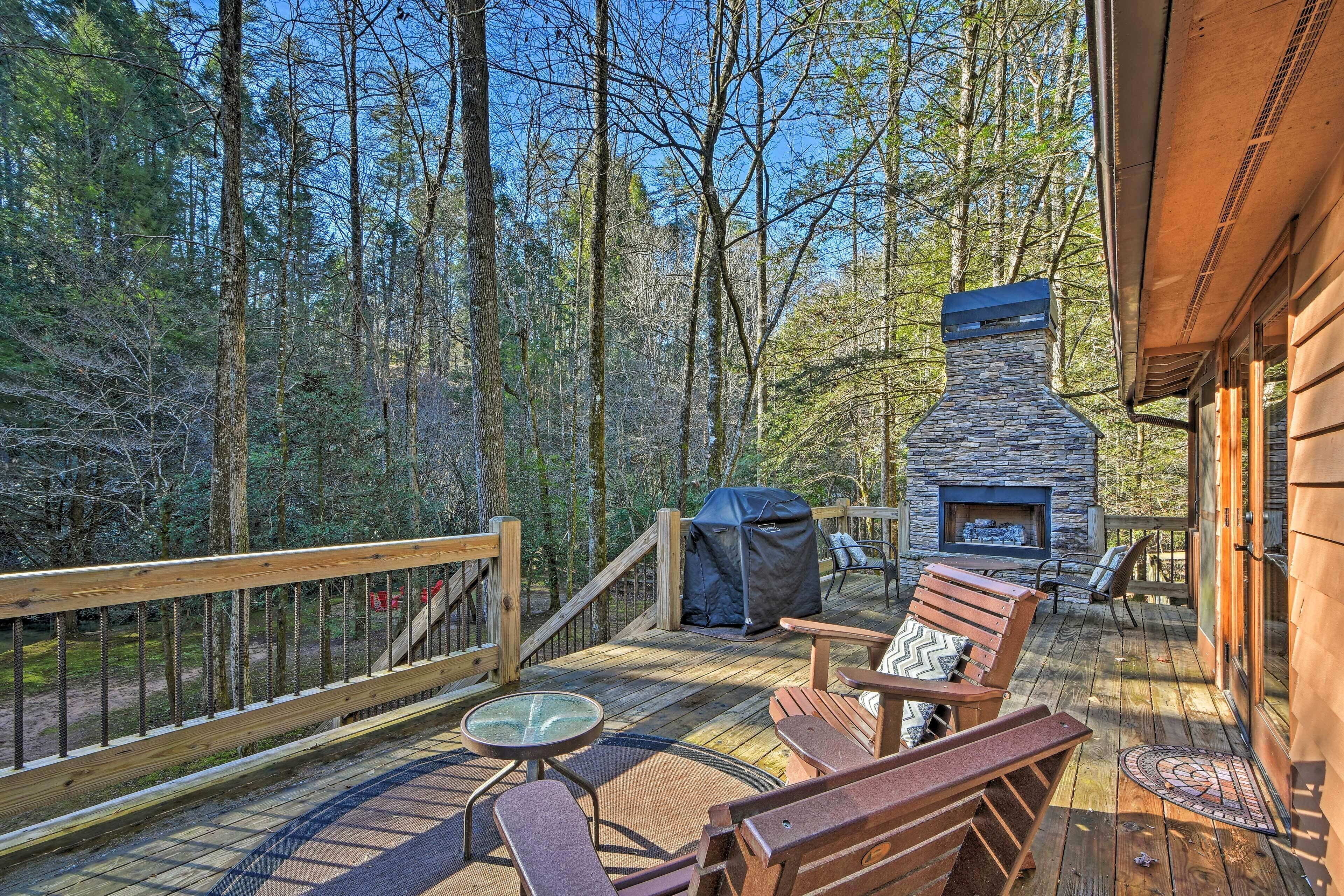 A wooden deck with outdoor furniture, a stone fireplace, and a view of the surrounding forest.