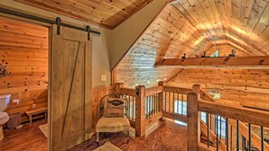 A wooden interior with a loft bedroom, a sliding barn door, a cushioned chair, and a view of the outside. - Blue Ridge Mtns Creekside Cabin w/ Hot Tub + Pier! (Blue Ridge)
