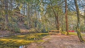 A forested area with a wooden bridge over a river, a picnic table, and red chairs. - Blue Ridge Mtns Creekside Cabin w/ Hot Tub + Pier! (Blue Ridge)