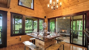 A wooden cabin interior with a large island table, benches, a fireplace, and large windows offering views of the outdoors. - Hot Tub + Views! Creekside Blue Ridge Mtn Escape (Mineral Bluff)