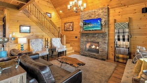 A cozy log cabin interior with a fireplace, wooden walls, a staircase, and comfortable seating. - Hot Tub + Views! Creekside Blue Ridge Mtn Escape (Mineral Bluff)