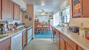 A kitchen with wooden cabinets, a dishwasher, a dining table with chairs, a ceiling fan, and a window with curtains.