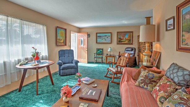 A cozy living room with a blue armchair, a rocking chair, a sofa with patterned cushions, a coffee table with books and a vase, and a door leading to another room.