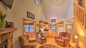 A cozy living room with a fireplace, plush armchairs, and a dining area with wooden cabinets and a kitchen in the background. - Modern Cabin w/ Deck in the Blue Ridge Mountains! (Blairsville)