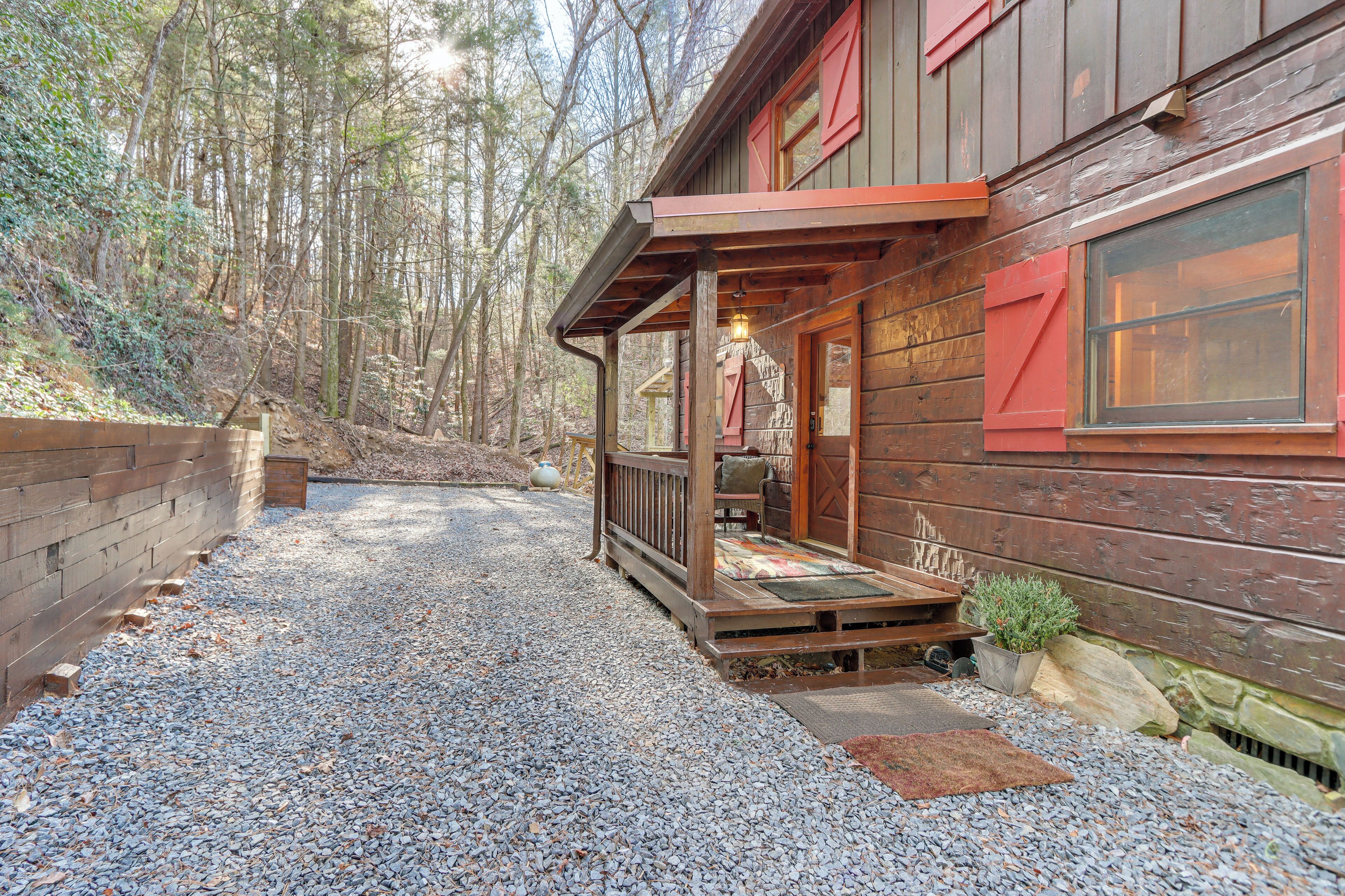 Une cabane en bois avec un porche, une allée en gravier et un paysage boisé en arrière-plan.