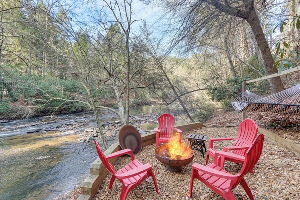 A fire pit with red chairs and a table in a forested area with a stream. - Waterfront Cabin w/ Beautiful River View & Hot Tub (Ellijay)