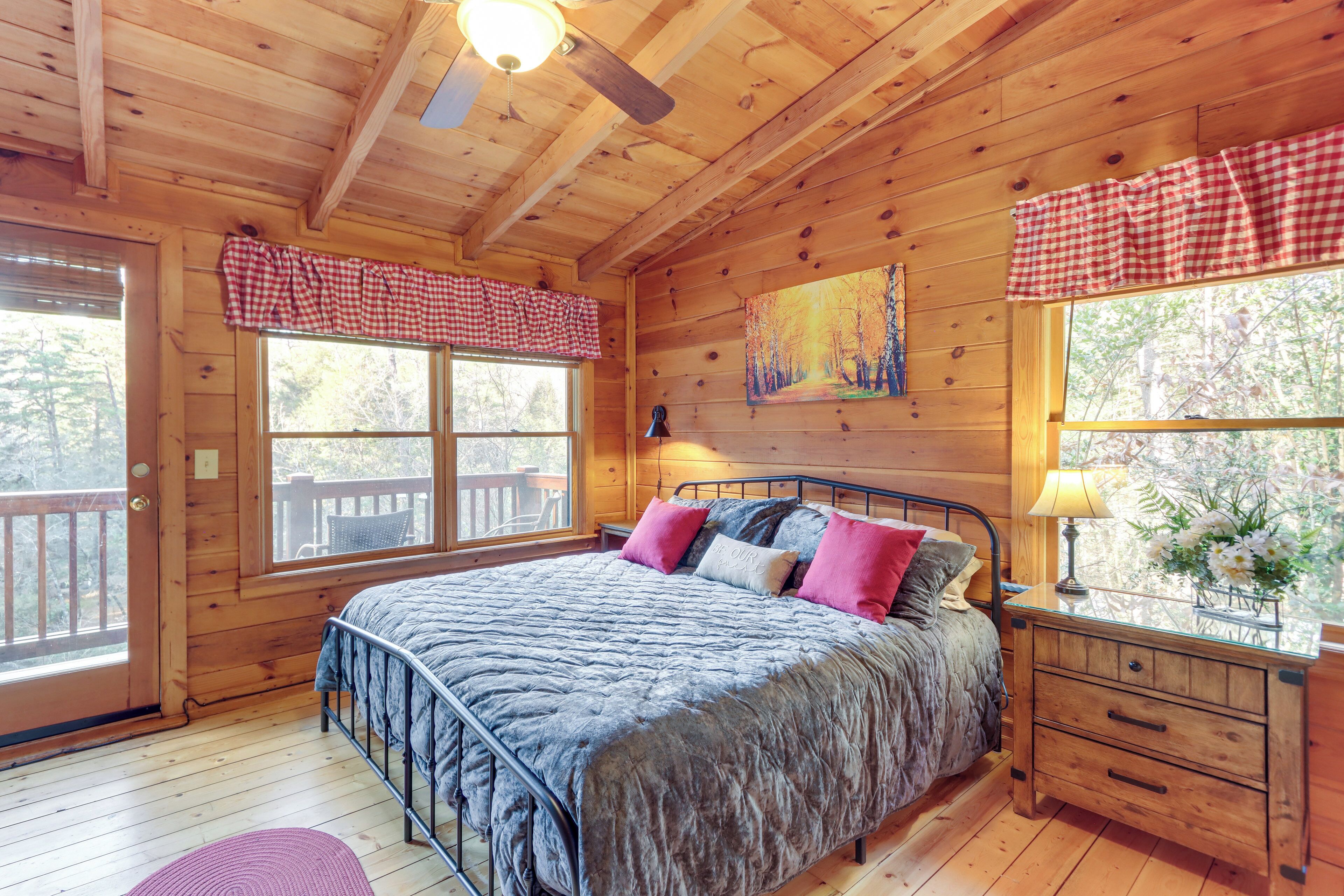 Un dormitorio de cabaña de madera con una cama, una mesita de noche, una ventana con un visillo a cuadros y un cuadro en la pared.