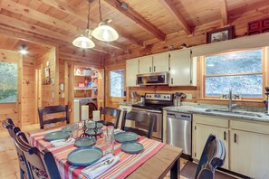 A wooden cabin interior with a dining table set for four, a kitchen with modern appliances, and a large window overlooking a snowy landscape. - Waterfront Cabin w/ Beautiful River View & Hot Tub (Ellijay)