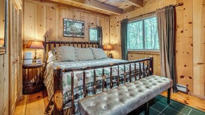 A wooden cabin bedroom with a bed, bedside lamps, a bench, and a window with curtains.