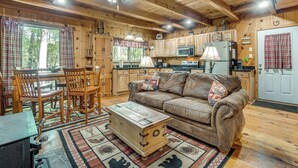 A rustic cabin interior with a wooden dining table, chairs, a sofa, and a kitchen area with wooden cabinets and appliances.