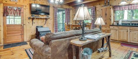 A cabin interior with a wooden ceiling, a television mounted above a fireplace, a sofa, a dining table, and a kitchen area.