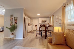 A modern living room with a beige sofa, a dining area with wooden chairs, a kitchen with white cabinets, and a hallway with a plant. - Hampton Vacation Rental: Central Location, Patio (Hampton)