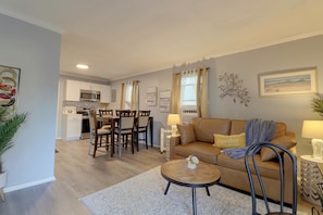 A living room with a sofa, a coffee table, and a dining area with chairs and a table. There is a kitchen in the background with cabinets and appliances. - Hampton Vacation Rental: Central Location, Patio (Hampton)