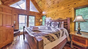 A wooden cabin bedroom with a large bed, a nightstand, and a window with blinds. - Unique Mineral Bluff Home With Mountain Views! (Mineral Bluff)
