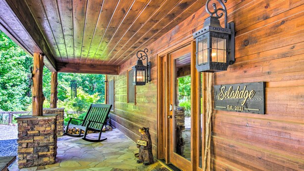 A wooden cabin entrance with a stone pillar, a welcome sign, a rocking chair, and a bear statue holding a sign. - Unique Mineral Bluff Home With Mountain Views! (Mineral Bluff)