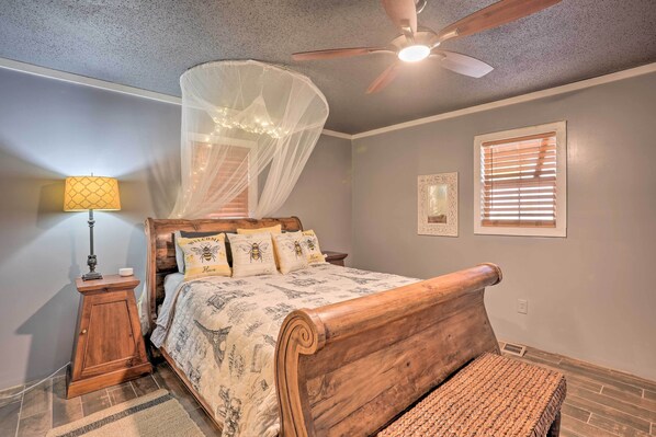 A bedroom with a wooden bed, a ceiling fan, a nightstand with a lamp, and a window with blinds. - Charming Escape w/ Fire Pit in Summerville! (Summerville)